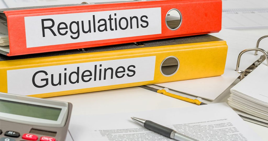 Red regulations binder and yellow guidelines binder on office desk with paperwork and pen for compliance documentation.